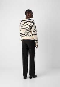 Gestrickter Pullover in Creme mit schwarzen abstrakten Mustern, kombiniert mit schwarzen Hosen. Ansicht von hinten, die das Design und die Passform des Pullovers hervorhebt.