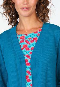Türkise Strickjacke mit strukturiertem Muster, getragen über einem blumigen Oberteil mit roten und pinken Blumen auf einem hellblauen Hintergrund.