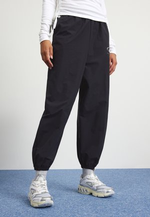 Personne portant un pantalon noir à poignets élastiques, un haut à manches longues blanc, des chaussettes grises et des baskets blanc-gris, debout sur un tapis bleu.