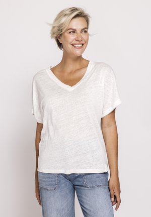 Lächelnde Frau mit kurzen blonden Haaren, die ein lockeres weißes T-Shirt mit V-Ausschnitt und hellblaue Jeans trägt, vor einem schlichten hellen Hintergrund stehend.