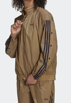Personne portant un survêtement beige Adidas avec des bandes noires et des détails à boutons, en train de fermer la veste d'une main.