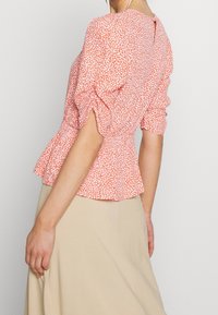 Peplum top s nabíranými rukávy, s bílým květinovým vzorem na korálové látce, texturovaný povrch a detail na zádech ve tvaru klíčové dírky.