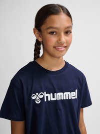 Młoda osoba z warkoczami, ubrana w granatowy T-shirt z białym logo „hummel” na jednolitym jasnoszarym tle.