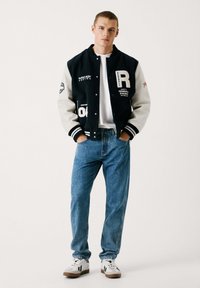 Chaqueta varsity negra y gris con logo "R", camiseta blanca, jeans azul relajados y zapatillas con patrón. Vestimenta deportiva casual.