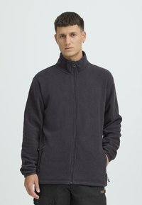 Tumehall fliisjakk kõrge kraega, täis tõmblukuga, küljetaskute ja pehme tekstuuriga. Rinnal minimaalne bränding.