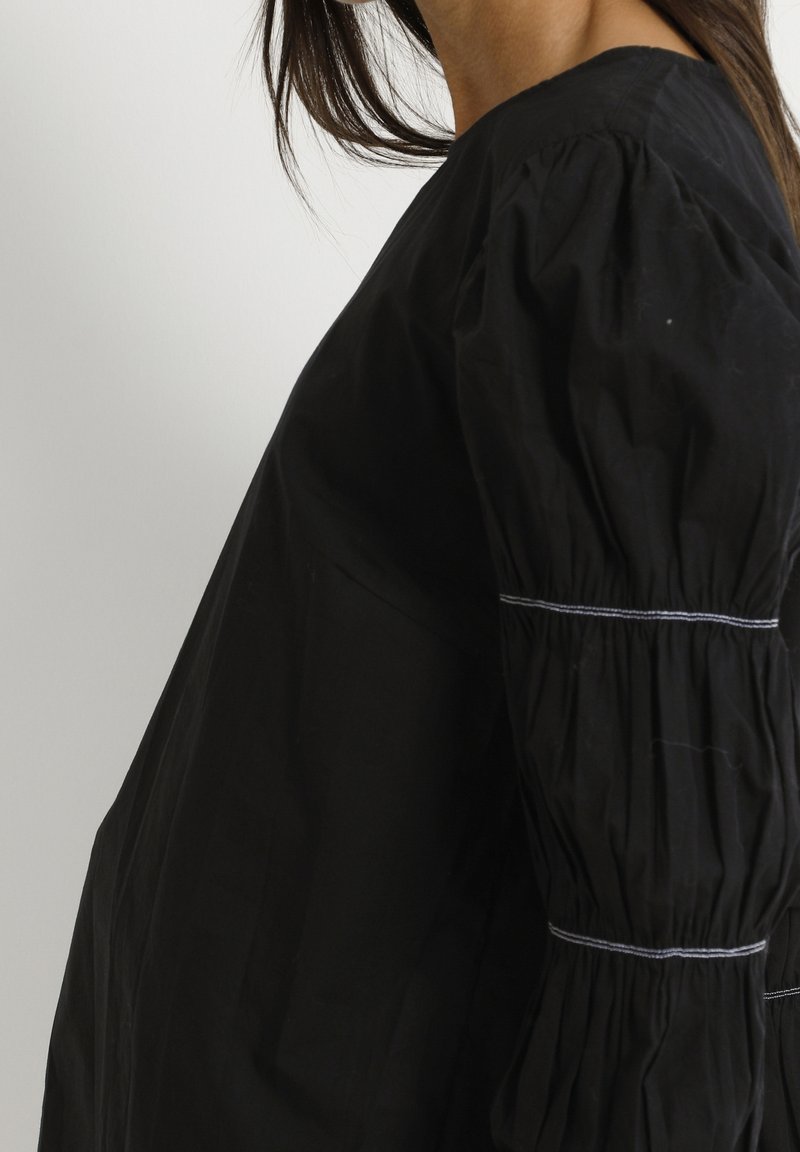 Femme portant un chemisier noir à manches bouffantes avec des détails blancs cousus sur le haut du bras et le bas de la manche.