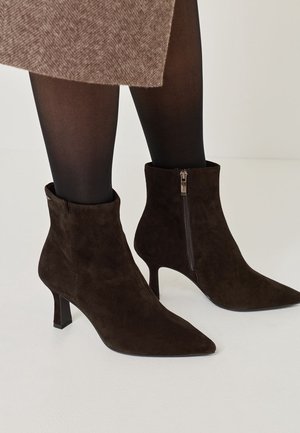 Femme portant des bottines en daim brun foncé à bouts pointus et fermetures éclair latérales, associées à des collants noirs et une jupe en tweed marron.