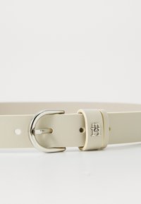 Beige nahkavyö, jossa on hopeanvärinen metallisolki, jossa on logoyksityiskohta ja useita säätöreikiä. Suuri sileä pinta ja minimalistinen muotoilu.