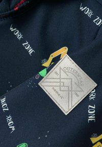 Ciemna tkanina z powtarzającym się napisem "STREFA ROBÓT", żółtymi ilustracjami koparek budowlanych oraz naszywaną kwadratową łatą z etykietą "ODZIEŻ WIERZCHNIA".