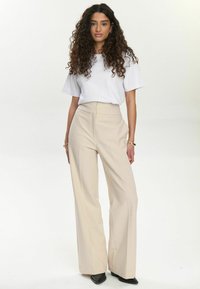 Pantaloni a vita alta e gamba larga in tessuto beige, abbinati a una camicia bianca a maniche corte. Tessuto morbido, design minimal e vestibilità su misura.