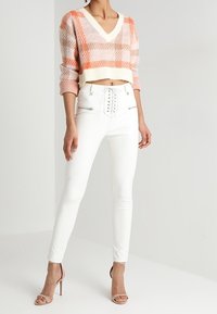 Maglione all'uncinetto in fantasia a scacchi arancione e bianco, scollatura a V; pantaloni bianchi aderenti con zip laterali; tacchi beige. Design minimalista.