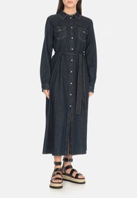 Abito camicia oversize in denim scuro con maniche lunghe, chiusura a bottoni, due tasche sul petto e cintura coordinata. Abbinato a sandali neri con platform.