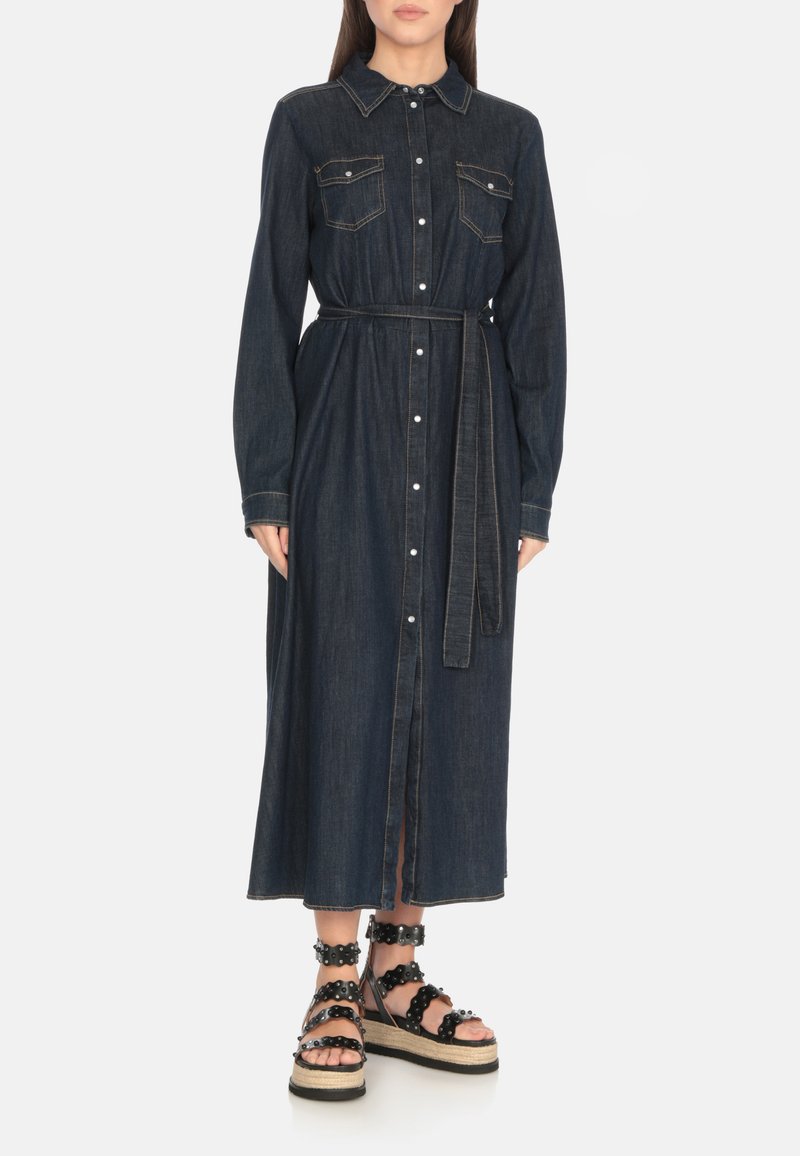 Abito camicia oversize in denim scuro con maniche lunghe, chiusura a bottoni, due tasche sul petto e cintura coordinata. Abbinato a sandali neri con platform.