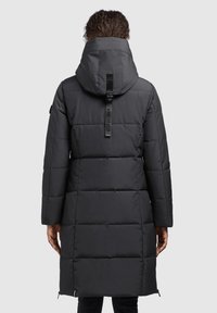 Woman wearing long black padded coat with hood and vertical strap detail on back, standing facing away against plain background.