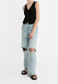 Czarna lniana kamizelka z dekoltem w serek i przednimi guzikami, zestawiona z jasnobłękitnymi spodniami typu wide-leg z przetarciami, które mają duże dziury na kolanach i postrzępione nogawki.