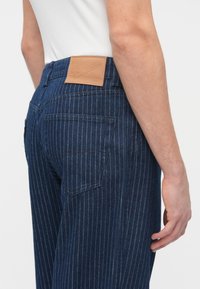 Τζιν παντελόνια με μοτίβο ρίγες, σκούρο μπλε χρώμα, δερμάτινο πίσω ετικέτα και πίσω τσέπες ορατές στην αριστερή πλευρά.