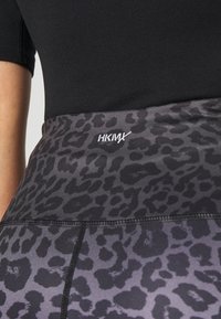 Zbliżenie na czarno-szary pasek w zwierzęcy wzór z białym logo "HKMX" na osobie noszącej czarną koszulkę.