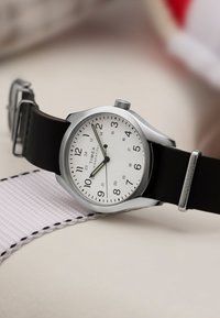Zilveren polshorloge met een wit wijzerplaat, zwarte rubberen band, duidelijke Arabische cijfers en een eenvoudig ontwerp met drie wijzers. Opvallend door de minimalistische stijl.
