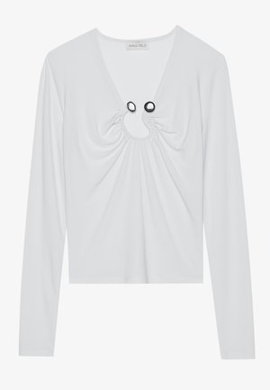 Biely dlhý rukáv top s hlbokým výstrihom do V, zdobený nariasenou látkou okolo výstrihu a dvomi čiernymi kruhovými akcentmi.