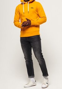Sudadera de felpa amarilla con cordones blancos, combinada con vaqueros de mezclilla oscura y zapatillas blancas con detalles en negro y lados texturizados.