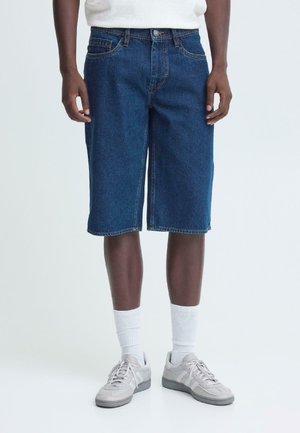 Donkerblauwe denim shorts met een losse pasvorm, voorzien van vijf zakken, riemlussen en gestikte zomen. Draagt witte sokken en grijze sneakers.