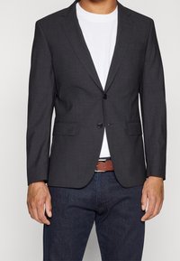 Ciemnoszary żakiet z dwoma przednimi kieszeniami i jednym zapięciem na guzik, noszony na białym t-shircie i niebieskich dżinsach. Gładka tekstura materiału.