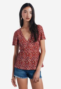 Rote Bluse mit Muster und kurzen Ärmeln, tiefem V-Ausschnitt und gerafftem Taillenbereich, kombiniert mit Jeansshorts. Sie hat geometrische weiße und orangefarbene Designs.