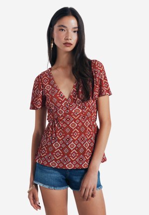 Rød mønstret bluse med korte ærmer, dyb V-hals og samlet talje, parret med denimshorts. Har geometriske hvide og orange mønstre.