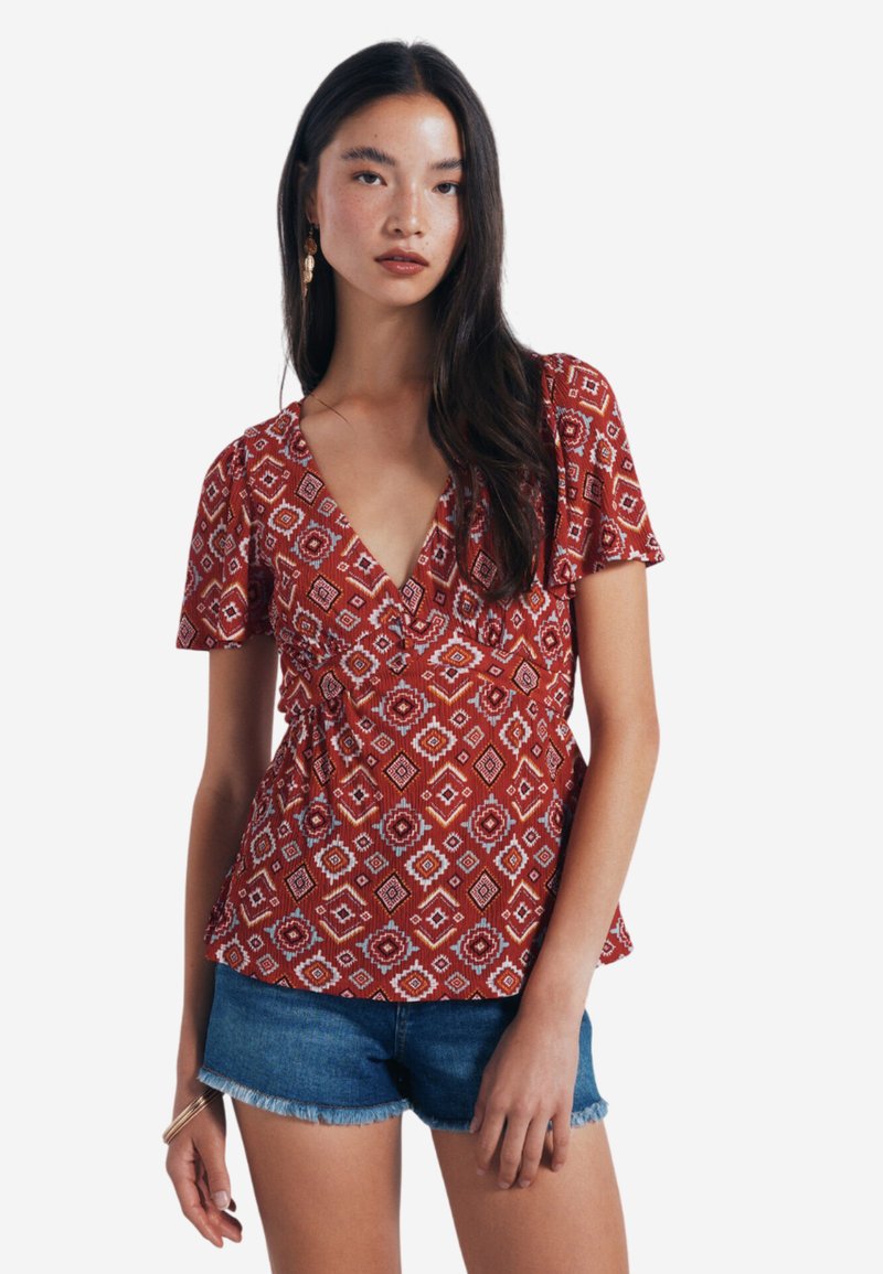 Rote Bluse mit Muster und kurzen Ärmeln, tiefem V-Ausschnitt und gerafftem Taillenbereich, kombiniert mit Jeansshorts. Sie hat geometrische weiße und orangefarbene Designs.