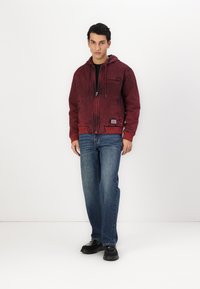 Rød hættetrøje med en tekstureret overflade, lynlås lukning og forlommer, sammen med blå jeans og sorte sko. Minimalistisk design, afslappet pasform.