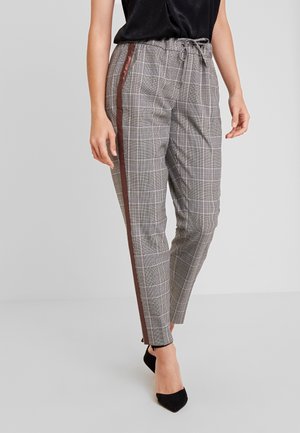 Kvinde iført grå plaid smalle bukser med brune sidelister, sort ærmeløs top og sorte spidse høje hæle, stående på gråt gulv.