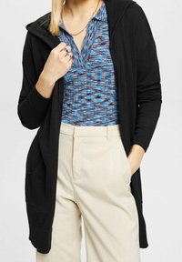 Svart, långärmad cardigan med huva, med två framfickor. Bärs över en blåmönstrad topp och krämfärgade byxor.