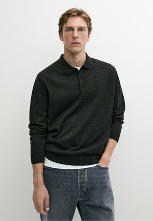 Junger Mann trägt ein dunkles langärmeliges Poloshirt über einem weißen Unterhemd und blauen Jeans, stehend vor einem schlichten weißen Hintergrund.