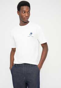 Λευκό βαμβακερό μπλουζάκι με μπλε γραφικό σχέδιο στην αριστερή πλευρά του στήθους, κοντά μανίκια και κλασικό στρογγυλό λαιμό. Συνδυασμένο με σκούρο τζιν παντελόνι.