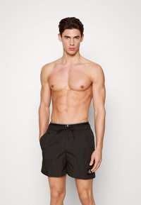 Shorts de bain noirs avec une taille élastique et un cordon de serrage, comprenant des poches latérales et un petit logo sur la jambe gauche. Texture de tissu lisse.