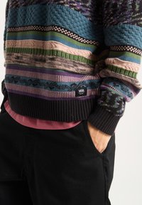 Multicolored knit sweater featuring textured patterns in blue, green, pink, and purple, with ribbed cuffs and a black logo tag on the hem.