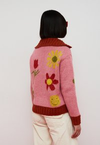 Knit sweater with a pink base, featuring floral patterns in red, yellow, and green; textured, with a ribbed orange collar and hem.