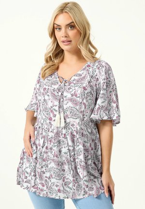 Kvinde iført en hvid bluse med pink paisley- og blomstermotiver, flagrende ærmer, kvastbånd ved halsudskæringen, kombineret med lyseblå jeans.