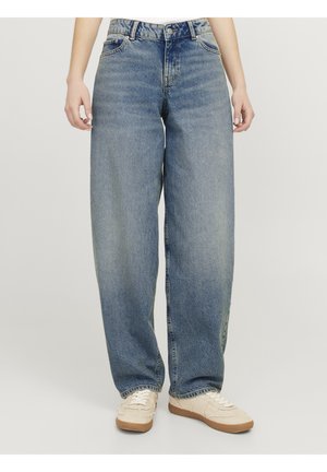 Person iført lyseblå vidde jeans og beige sneakers, der står mod en ensfarvet lys baggrund.