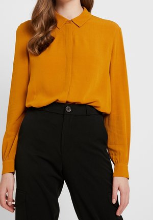 Woman wearing a mustard yellow long-sleeve blouse tucked into high-waisted black pants with a button closure.