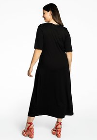 Femme portant une robe midi noire avec des manches coude, se tenant debout et regardant sur le côté, associée à des sandales à talons hauts à motifs rouge et blanc.