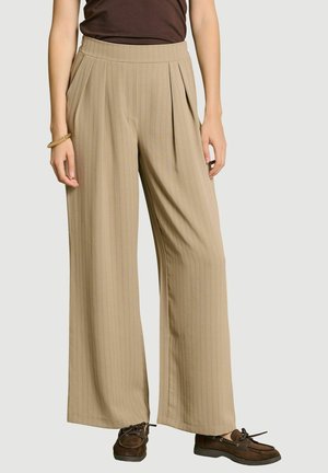 Femme portant un pantalon beige large plissé, un haut marron foncé, des mocassins marron et un bracelet en or au poignet gauche.