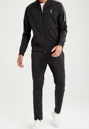 Mand i sort lynlåstrøje og sorte bukser går, iført hvide sneakers mod en ensfarvet lys baggrund.