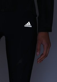 Zwarte sportleggingen met een reflecterend wit Adidas-logo op de dij. Close-up van een hand die op de stofstructuur rust.