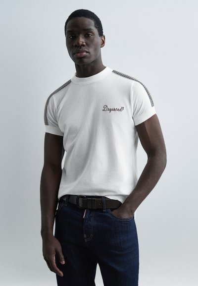 Homme portant un t-shirt blanc Dsquared2 avec des détails rayés sur les épaules et un jean bleu foncé, debout avec une main dans la poche.