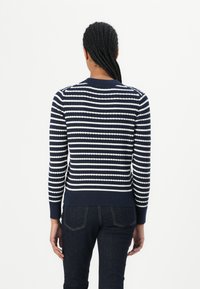 Marineblauwe trui met witte horizontale strepen, ribbeltextuur en aansluitend ontwerp. Heeft een ronde hals en lange mouwen, gecombineerd met donkere jeans.