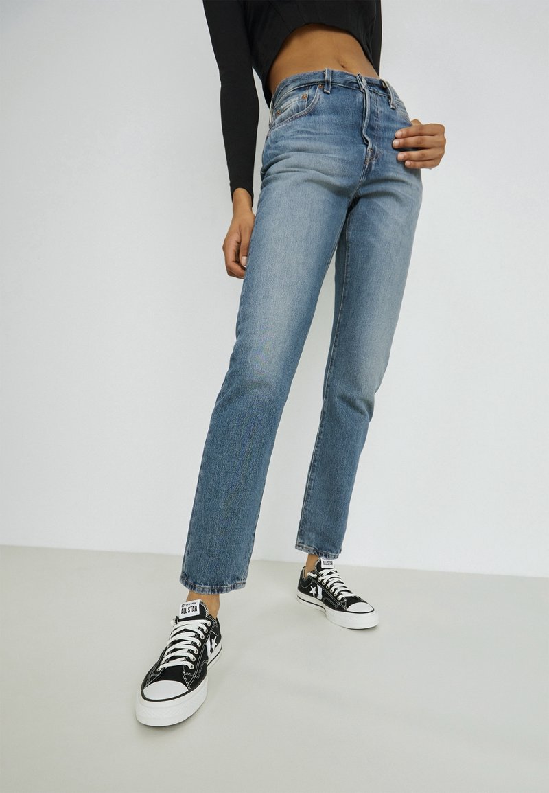 Personne portant un jean bleu taille haute, un crop top noir à manches longues et des baskets style Converse noir et blanc, debout sur un sol uni.