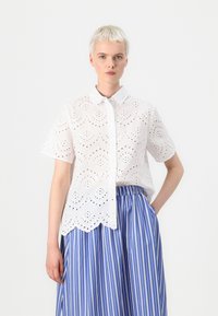 White eyelet shirt with short sleeves, button-up front, scalloped hem, and cut-out patterns. Paired with a blue striped skirt.