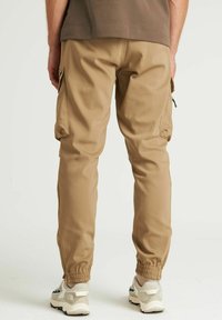 Beige cargo broek met elastische boorden, zijzakken en een gladde stofstructuur. Draagt met lichtgekleurde sneakers.