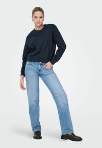 Sweatshirt noir à col rond et manches longues, associé à un jean droit bleu clair. Le modèle se tient avec assurance, les mains sur les hanches.